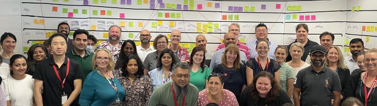 A OneCX Program staff group photo in front of a whiteboard with colourful sticky notes and writing on it.